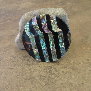 May include: A round, black and iridescent abalone shell brooch with a geometric pattern of vertical stripes.