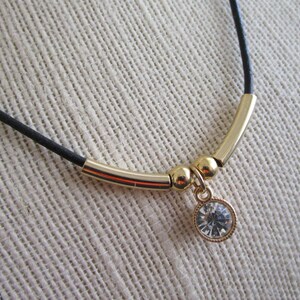 May include: A gold necklace with a black cord and a single crystal pendant. The pendant is round and has a faceted design.