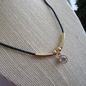 May include: A black leather cord necklace with a gold bar and small gold beads. A clear crystal pendant hangs from the bar.
