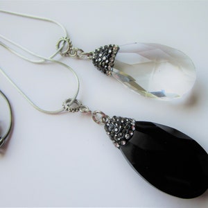 May include: Two teardrop-shaped pendant necklaces. One pendant is clear, the other is black. Both pendants have a silver-colored metal cap encrusted with small, clear crystals. The necklaces have a silver-colored chain.