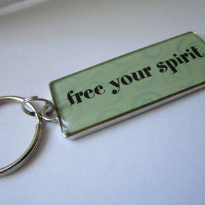 Puede incluir: Un llavero de plata con una etiqueta rectangular que dice "libera tu espíritu" en texto negro sobre un fondo verde claro con un patrón de espiral.