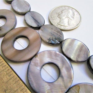 May include: A strand of oval and round shell beads with an iridescent finish. The beads are a mix of light and dark grey with a brown undertone. The beads are strung on a white thread. A UK 20 pence coin is included for size reference.