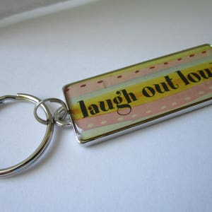 Puede incluir: Un llavero de plata con un dije rectangular que dice "laugh out loud" en texto negro sobre un fondo rayado amarillo, rosa y blanco.