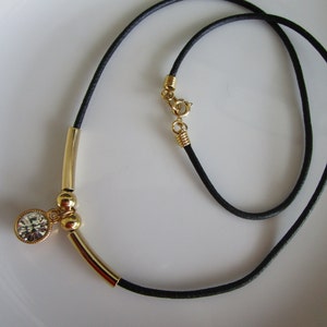 May include: A gold necklace with a black cord and a single crystal pendant. The pendant is attached to a gold bar with two gold beads.