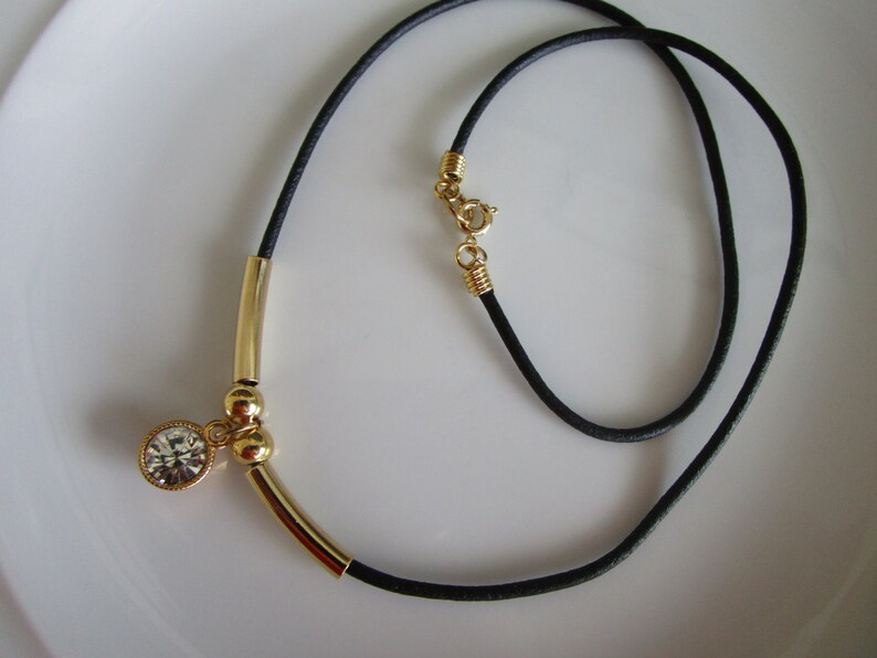 May include: A gold necklace with a black cord and a single crystal pendant. The pendant is attached to a gold bar with two gold beads.