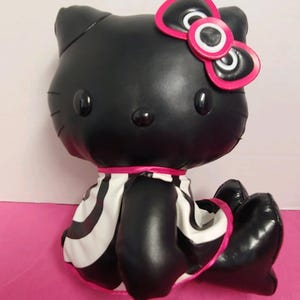 May include: A black plush cat figure with a pink bow and a black and white striped skirt. The cat has black eyes and a pink ribbon around its neck. The figure is sitting on a pink surface.