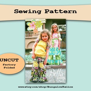 May include: A sewing pattern image featuring two young girls wearing colorful floral pants and tops. The text "Sewing Pattern" is at the top, with "UNCUT Factory Folded" in an oval. The pattern is for "the Bella pants No. 2".