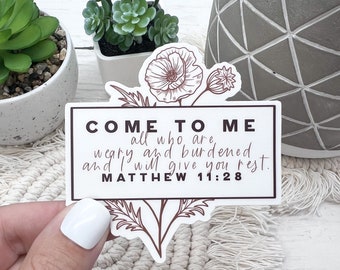 May include: A rectangular sticker with the text "COME TO ME all who are weary and burdened and I will give you rest. MATTHEW 11:28." The sticker has a floral design in brown and is held by a hand.