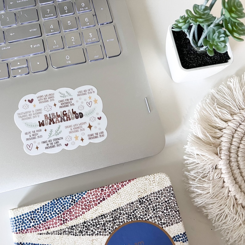 May include: A sticker with a brown cloud shape and white text that reads "Biblical Womanhood" with bible verses and other words in a brown font. The sticker is on a silver laptop.
