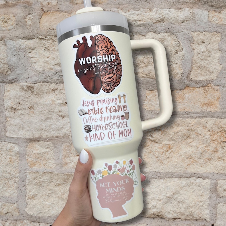 Puede incluir: Una taza de viaje blanca con una pegatina rosa y marr&oacute;n que dice "Worship in spirit and truth" y una lista de actividades que incluyen "Jesus praising, Bible reading, Coffee drinking, Homeschool, *Kind of Mom". La taza tambi&eacute;n tiene una pegatina con un dise&ntilde;o floral y el texto "Set your minds on things above, not on earthly things." Filipenses 4:8.