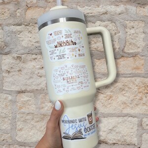 May include: A white tumbler with a handle, featuring several stickers with bible verses and quotes about love and womanhood. The tumbler is decorated with hearts and floral designs. The text on the stickers includes "Biblical Womanhood", "Biblical Love", and "Mornings with Jesus Coffee are my favorite".