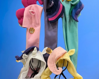 May include: Five fleece hooded scarves featuring character designs with ears and horns. The collection includes a pink scarf with a curly mane, a purple one with a dark horn, a teal scarf with a rainbow mane, a white one with a horn, and a yellow scarf.
