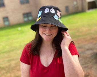 May include: A black bucket hat featuring a pair of white cartoon eyes on the front and various colorful, star-shaped and cloud-shaped patches attached around the crown. The hat has a wide, downward-sloping brim and a flat top construction.
