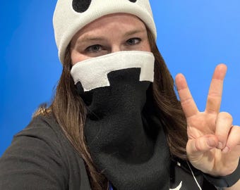May include: A white knit beanie featuring two black oval eye shapes and a matching white and black face mask covering the nose and mouth. The mask has a jagged, tooth-like design across the top edge, with a solid black lower section.