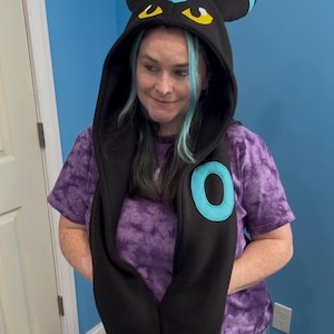 May include: Black hooded scarf with a cartoon-style design featuring blue and yellow accents. The scarf has a large blue circle on the front and is paired with a purple tie-dye t-shirt and ripped jeans.