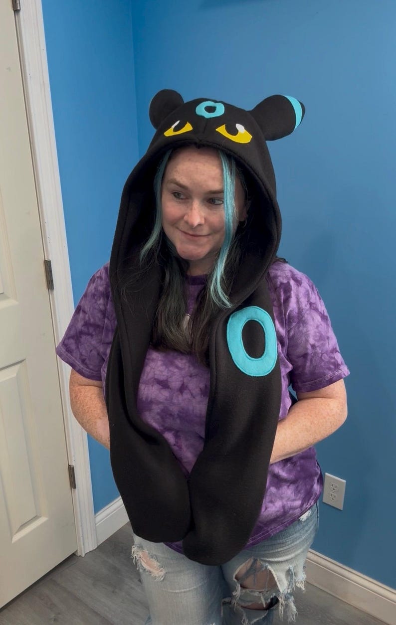 May include: Black hooded scarf with a cartoon-style design featuring blue and yellow accents. The scarf has a large blue circle on the front and is paired with a purple tie-dye t-shirt and ripped jeans.
