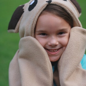 May include: A beige hooded hat with attached mittens, designed to resemble a cartoon animal. The hat features large black eyes, brown ears, and a spiky crown. The mittens are attached to the hood, framing the face. The person is wearing a blue shirt.