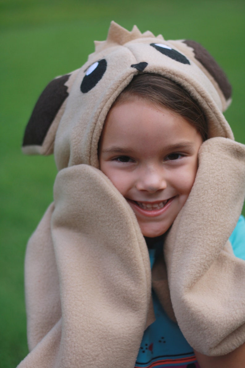 May include: A beige hooded hat with attached mittens, designed to resemble a cartoon animal. The hat features large black eyes, brown ears, and a spiky crown. The mittens are attached to the hood, framing the face. The person is wearing a blue shirt.