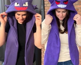 May include: A purple hooded scarf featuring a cartoon character face with white eyes, a red mouth, and pointed ears. The scarf is made of a soft material and has long, draping sides. The hood is up, and the scarf is being worn.