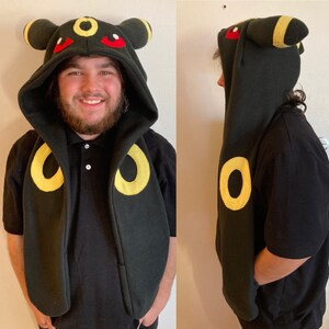 May include: Black hooded cloak with yellow and red accents, resembling a character from a popular animated series. The cloak features large yellow circles and a hood with ears and red eyes. The design is visible from the front and side.