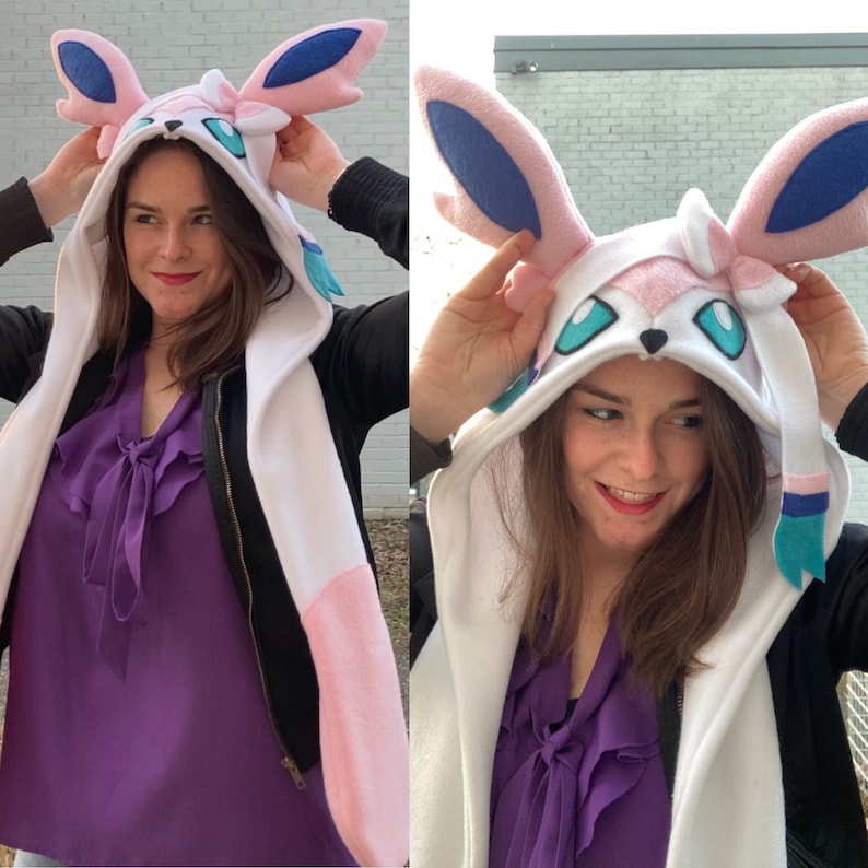 May include: A white hooded scarf with pink and blue accents, resembling a Sylveon character. The hood features large pink ears with blue inner details and teal accents. The scarf is white with pink details.