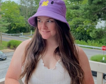 May include: A purple bucket hat with a yellow cross and black dot design. The hat is made of a soft material and has a wide brim. The hat is worn by a person.