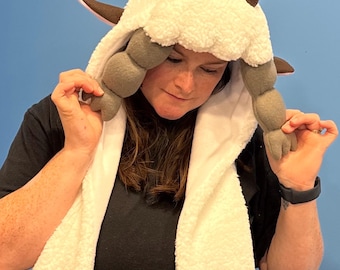 May include: A white hooded scarf with a plush texture, featuring brown horns and ear details. The scarf has gray, segmented appendages. The design resembles a character from a popular animated series. The background is a solid blue.