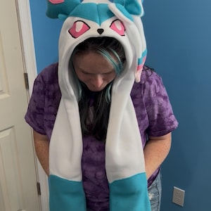 May include: A hooded hat with long scarf-like extensions, featuring a Sylveon design. The hat is primarily white with accents of light blue, pink, and black. The ears and face details are prominent, creating a playful, character-inspired accessory.