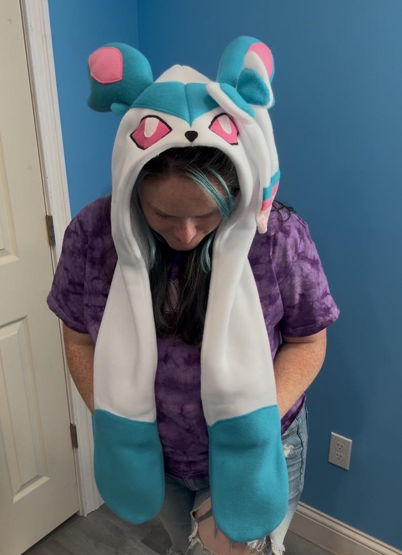 May include: A hooded hat with long scarf-like extensions, featuring a Sylveon design. The hat is primarily white with accents of light blue, pink, and black. The ears and face details are prominent, creating a playful, character-inspired accessory.