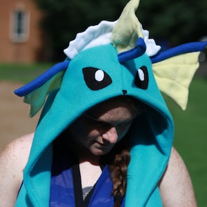 May include: A teal hooded sweatshirt with a Vaporeon design. The hood features black eyes, blue accents, and yellow fins. The sweatshirt is made of soft fabric and is designed to resemble the Pok&eacute;mon character.