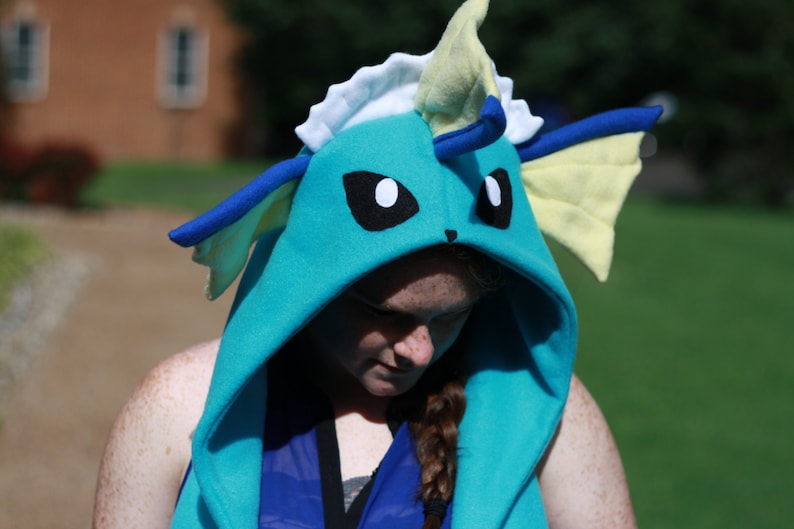 May include: A teal hooded sweatshirt with a Vaporeon design. The hood features black eyes, blue accents, and yellow fins. The sweatshirt is made of soft fabric and is designed to resemble the Pok&eacute;mon character.
