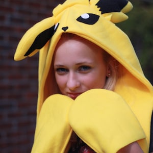 May include: A yellow Pikachu costume featuring a hooded sweatshirt with black ears and facial details, and matching yellow mittens. The costume is worn by a person, with a brick wall in the background.
