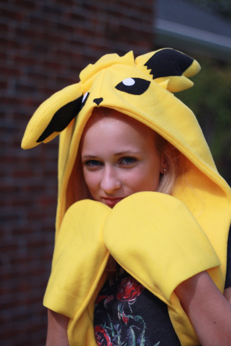 May include: A yellow Pikachu costume featuring a hooded sweatshirt with black ears and facial details, and matching yellow mittens. The costume is worn by a person, with a brick wall in the background.