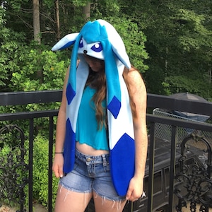 May include: A blue and white hooded scarf resembling a cartoon character. The scarf has long, dangling blue accents and large, pointed ears. The hood features a face with large purple eyes. The scarf is worn over a teal top and denim shorts.