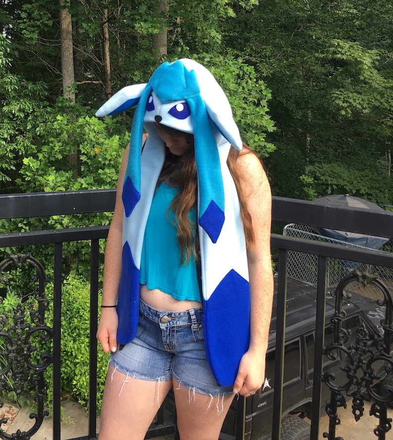 May include: A blue and white hooded scarf resembling a cartoon character. The scarf has long, dangling blue accents and large, pointed ears. The hood features a face with large purple eyes. The scarf is worn over a teal top and denim shorts.