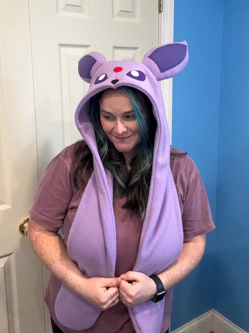 May include: A lavender fleece hood with large purple ears and a red dot on the forehead. The hood has long scarf-like extensions. The hood is designed to resemble a character from a popular animated series.