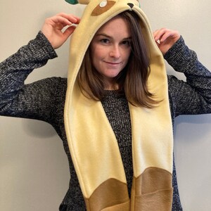 May include: A beige and brown hooded scarf with a cartoon animal design. The hood features ears and facial details, with the scarf extending down the front. The scarf has pockets at the bottom. The person in the image is wearing a dark gray sweater.