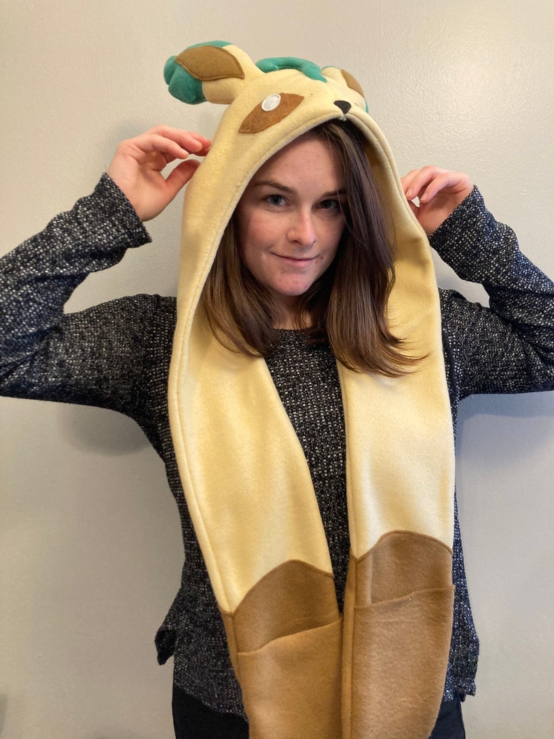 May include: A beige and brown hooded scarf with a cartoon animal design. The hood features ears and facial details, with the scarf extending down the front. The scarf has pockets at the bottom. The person in the image is wearing a dark gray sweater.