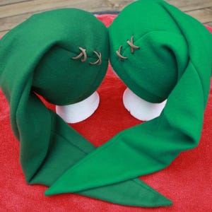 May include: Two green elf hats with long, pointed tips. Each hat has a brown leather-like accent stitched on the side. The hats are displayed on white forms against a red background.