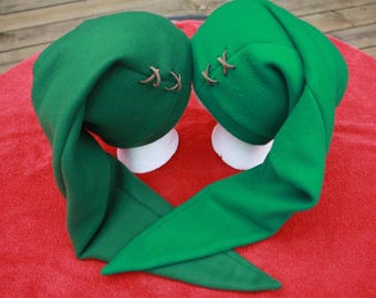 May include: Two green elf hats with long, pointed tips. Each hat has a brown leather-like accent stitched on the side. The hats are displayed on white forms against a red background.