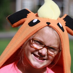 May include: An orange hooded costume resembling a cartoon character with black ears, white eyes, and a yellow flame-shaped top. The costume is worn by a person wearing glasses and a pink shirt, smiling at the camera.