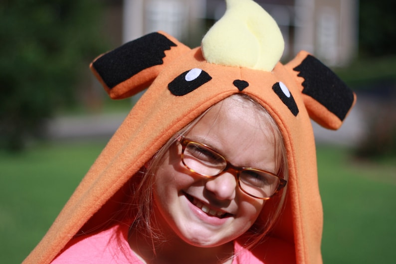 May include: An orange hooded costume resembling a cartoon character with black ears, white eyes, and a yellow flame-shaped top. The costume is worn by a person wearing glasses and a pink shirt, smiling at the camera.