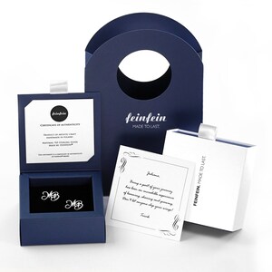 May include: A blue jewelry box with a pair of silver earrings inside. The earrings are initial earrings with the letters "MB". The box is open and the earrings are visible. The box is on a white surface. The box has a certificate of authenticity inside. The box is also accompanied by a white card with a handwritten message. The card is on a white surface. The box is also accompanied by a blue shopping bag with the word "feinfein" printed on it. The bag is on a white surface.