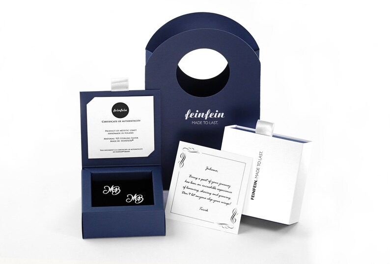 May include: A blue jewelry box with a pair of silver earrings inside. The earrings are initial earrings with the letters "MB". The box is open and the earrings are visible. The box is on a white surface. The box has a certificate of authenticity inside. The box is also accompanied by a white card with a handwritten message. The card is on a white surface. The box is also accompanied by a blue shopping bag with the word "feinfein" printed on it. The bag is on a white surface.