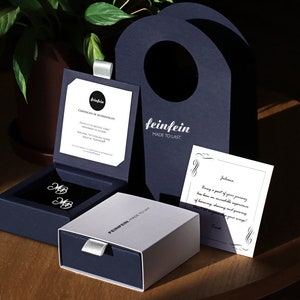 May include: A white box with the text "feinfein made to last" on it, next to a blue box with two silver cufflinks inside. The cufflinks are in the shape of the letter "MB". There is a white card with a handwritten message on it, and a blue bag with the text "feinfein made to last" on it.