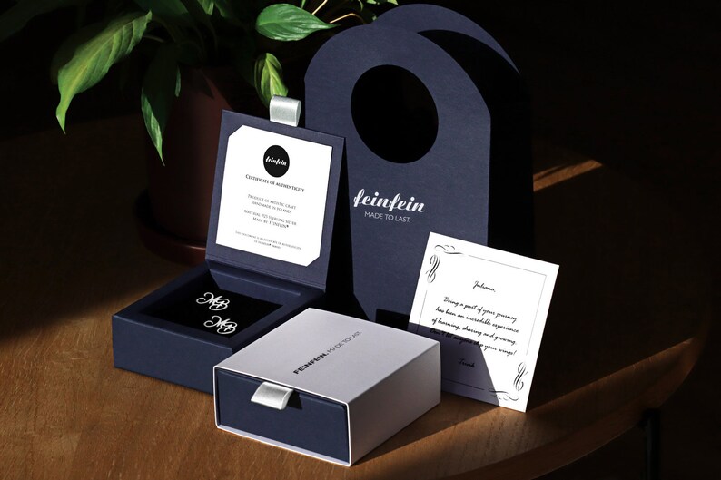 May include: A white box with the text "feinfein made to last" on it, next to a blue box with two silver cufflinks inside. The cufflinks are in the shape of the letter "MB". There is a white card with a handwritten message on it, and a blue bag with the text "feinfein made to last" on it.