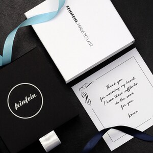 May include: A black gift box with a white circle logo that says "feinfein" on it. The box is tied with a white ribbon. A white card with black text says "Thank you for warming my heart. I hope these cufflinks do the same for you. Karen."