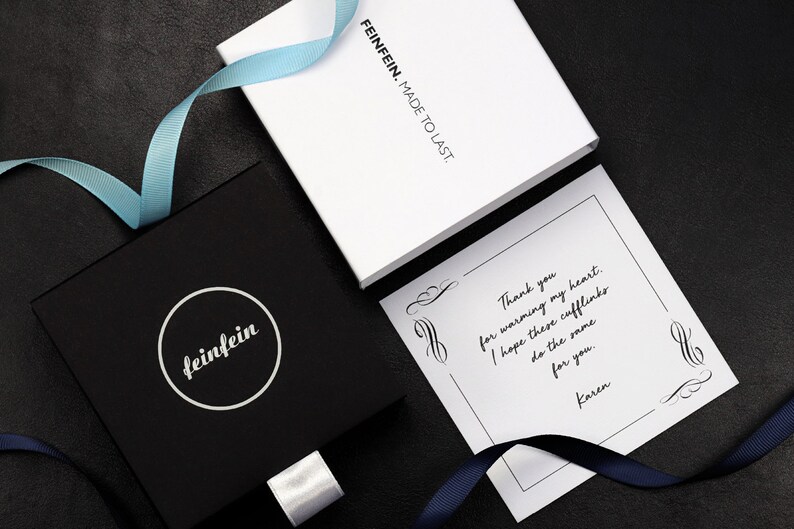 May include: A black gift box with a white circle logo that says "feinfein" on it. The box is tied with a white ribbon. A white card with black text says "Thank you for warming my heart. I hope these cufflinks do the same for you. Karen."