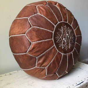 K&ouml;nnte beinhalten: Runder brauner Leder-Ottoman mit silbernen N&auml;hten und einem Schneeflocken-Design in der Mitte. Der Ottoman ist ein dekoratives M&ouml;belst&uuml;ck, perfekt f&uuml;r ein Wohn- oder Schlafzimmer. Der Ottoman hat einen Durchmesser von ca. 51 cm.