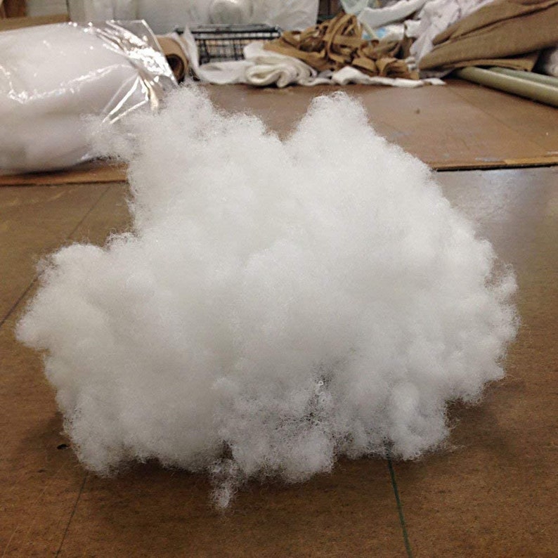 Op de afbeelding: Een grote, pluizige wolk van witte polyester vulling, ideaal voor het vullen van kussens, kussens of knutselprojecten. De zachte, wolkenachtige textuur is zichtbaar in deze close-up. De vulling wordt getoond op een bruin oppervlak.